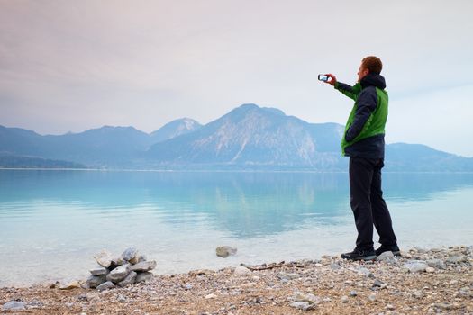 Tall man hold cellphone, take picture of autumn mountain lake scenery after rainy day. Vivid and vignetting effect. Poor lighting conditions.