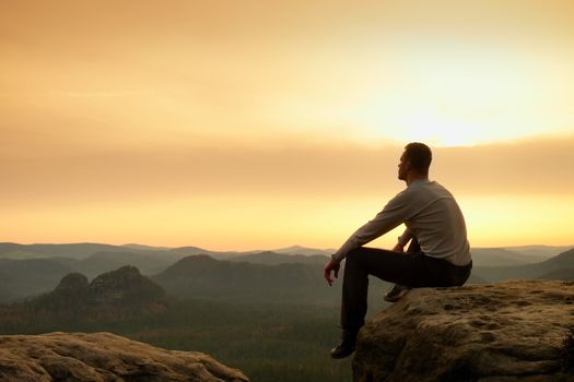 Hiker in black sit alone on the rock summit. Wonderful daybreak in mountains, heavy mist in deep valley. Man sit on the rock.