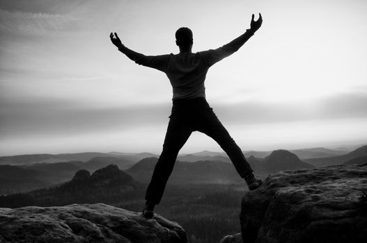 Happy man with raised arms gesture triumph on exposed cliff. Satisfy hiker silhouette on sandstone cliff watching down to hilly landscape.