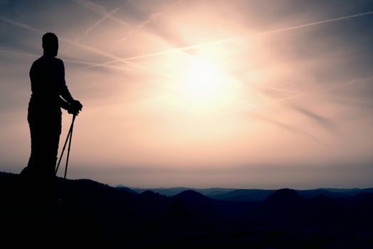 Silhouette of tourist with poles in hand. Sunny spring daybreak in rocky mountains. Hiker w on rocky view point above misty valley. 