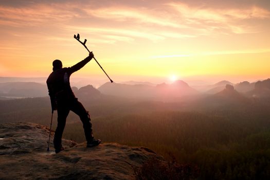 Happy man with broken leg in immobilizer and medicine pole above head stay on peak. Open misty mountain valley bellow cliff. Silhouette of tourist with hand in air. Spring daybreak