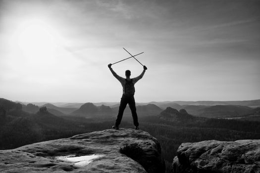 Happy backpacker with x crossed poles in the air, open misty mountain valley bellow cliff. Silhouette of tourist with poles in hands. Sunny spring daybreak in hills.