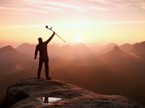Hiker with  medicine crutch above head achieved mountain peak. Broken leg fixed in immobilizer Deep misty valley bellow silhouette of man with hand in air. Spring daybreak