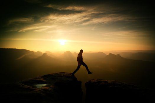 Crazy hiker in black is jumping between rocky peaks. Happy man. Wonderful daybreak in rocky mountains, heavy orange mist in deep valley. Miracle of nature