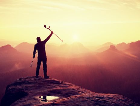 Happy man with broken leg and medicine crutch.  Hiker with leg in immobilizer achieve peak of mountain. Daybreak misty valley bellow silhouette. 