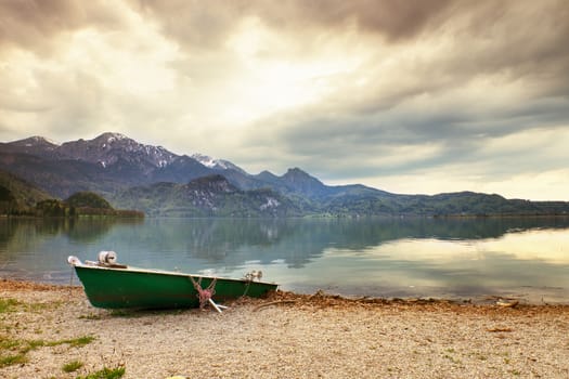 Abandoned fishing paddle boat on bank of Alps lake. Morning lake glowing by sunlight. Dramatic and picturesque scene. Mountains in water mirror.