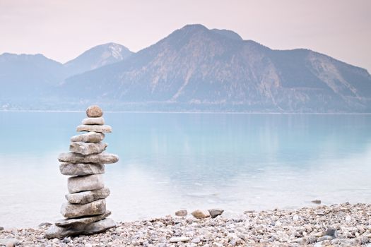 Balanced stone pyramide on shore of blue water of mountain lake. Blue mountains in water level mirror. Children built pyramid from pebbles.  Poor lighting conditions.