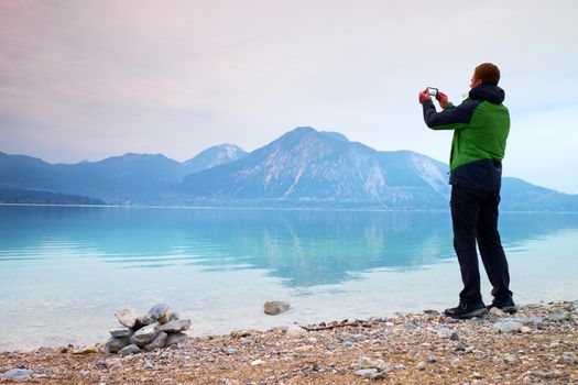 Tall man hold cellphone, take picture of autumn mountain lake scenery after rainy day. Vivid and vignetting effect. Poor lighting conditions.