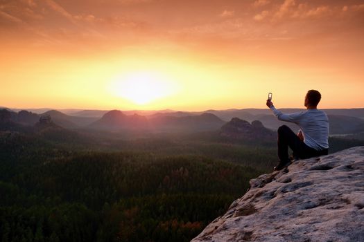 Tourist in grey t-shirt takes photos with smart phone on peak of rock. Dreamy hilly landscape below, spring orange pink misty sunrise in beautiful valley below rocky mountains. Hiking man taking photo