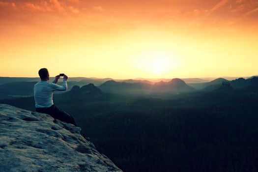 Tourist in grey t-shirt takes photos with smart phone on peak of rock. Dreamy hilly landscape below, spring orange pink misty sunrise in beautiful valley below rocky mountains. Hiking man taking photo