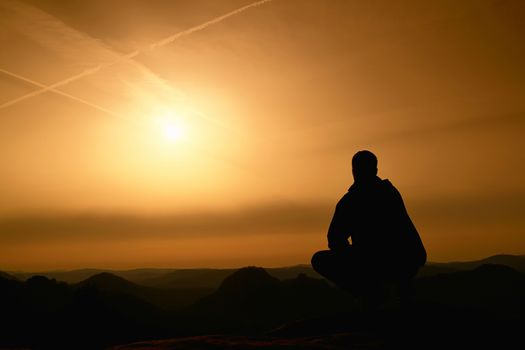 Adult hiker in black sit on mountain  edge. Man enjoying evening and looking to orange misty horizon