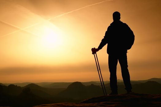 Tall backpacker with poles in hand. Sunny spring daybreak in rocky mountains. Hiker with big backpack stand on rocky view point above misty valley. 