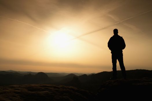 Sportsman in black with hands in pockets stand on the peak in rock empires park and watching over the misty and foggy morning valley to Sun. Silhouette of man.