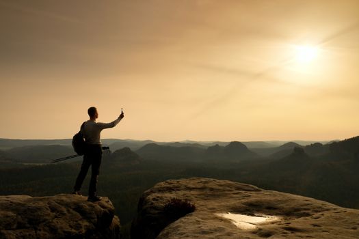 Tall tourist at dangerous cliff edge is taking selfie on peak above valley. Mountain peak and deep valley with sun in the frame