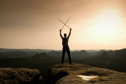 Happy backpacker with x crossed poles in the air, open misty mountain valley bellow cliff. Silhouette of tourist with poles in hands. Sunny spring daybreak in hills.