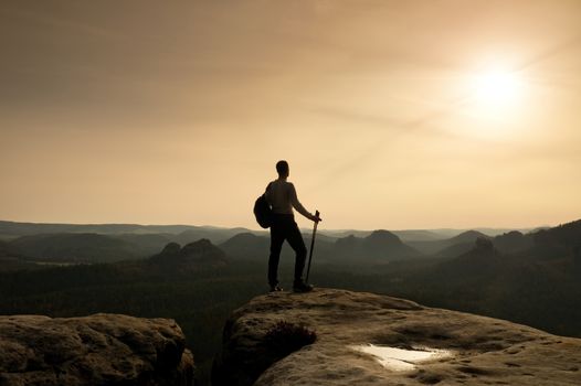 Tourist guide on cliff edge with pole in hand. Hiker with sporty backpack stand on rocky view point above misty valley. Sunny spring daybreak in rocky mountains.