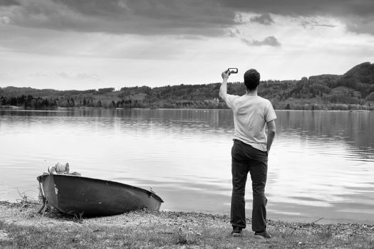 Man in blue shirt take photo by smart phone. Silhouette at fishing paddle boat at mountains lake coast.