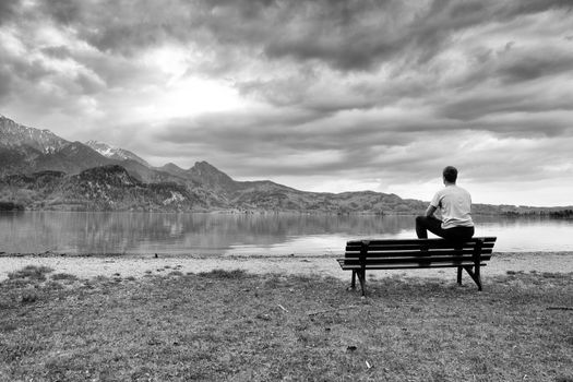 Man take rest. Wooden bench for relaxing on shore. View on Alps mountains with white peaks mirror in lake level.