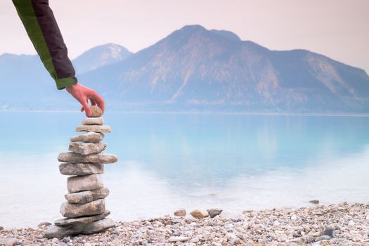 Man built pyramid from pebbles. Balanced stone pyramide on shore of blue water of mountain lake. Blue mountains in water level mirror.  Poor lighting conditions.