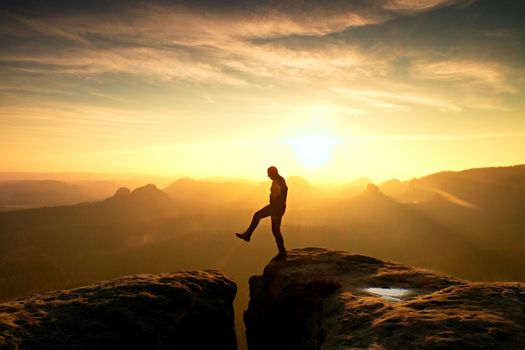 Man in black jumping between rocky empires. Dreamy daybreak in sandstone mountains, heavy orange mist in deep valley.