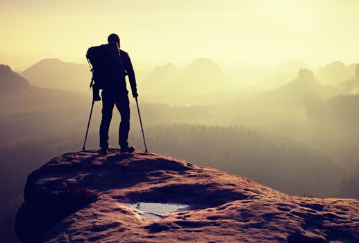 Tourist with medicine crutch above head achieved mountain peak. Hiker with broken leg in immobilizer. Deep misty valley bellow silhouette of man with hand in air. Spring daybreak