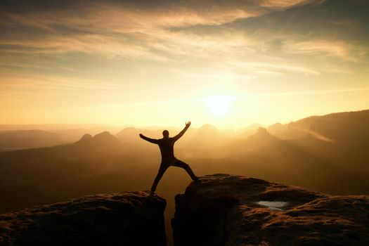 Hiker in black celebrate triumph between two rocky peaks. Wonderful daybreak in rocky mountains, heavy orange mist in deep valley