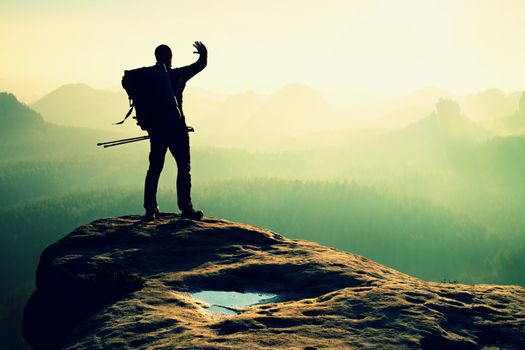 Hiker on peak. Backpacker with poles in hand shadowing eyes. Sunny spring daybreak in rocky mountains. Hiker on rocky view point above misty valley. 