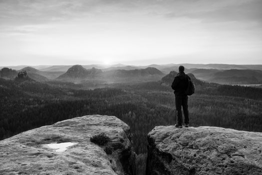 Tall tourist guide on rock with pole in hand. Hiker with sporty backpack stand on cliff above misty valley. Sunny spring daybreak in rocky mountains.