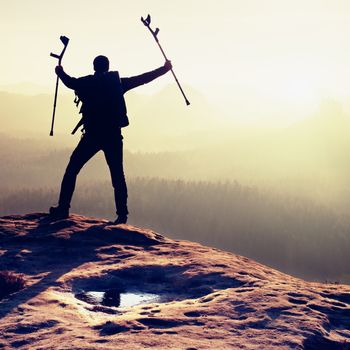 Tourist with  medicine crutch above head achieved mountain peak. Hiker with broken leg in immobilizer.  Deep misty valley bellow silhouette of man with hand in air. Spring daybreak