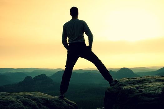 Sportsman in black with hands in pockets stand on the peak in rock empires park and watching over the misty and foggy morning valley to Sun. Silhouette of man.