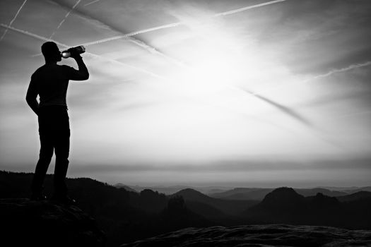 Thirsty hiker in black pants with bottle of water. Sweaty tired tourist on the peak of sandstone rocky park Saxony Switzerland watching into misty landscape.