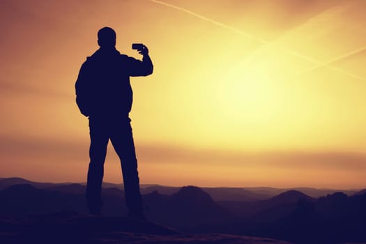 Tall hiker hold in one hand mobile phone and  take photo of misty landscape. Smart phone photography on  the peak of mountain at sunrise.