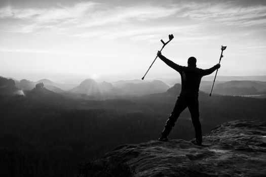 Tourist with forearm crutch above head achieved mountain peak. Hiker with broken leg in immobilizer and medicine  poles hold hand in air. Colorful misty valley bellow silhouette.