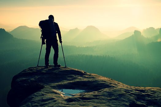 Tourist with leg in immobilizer. Hiker silhouette with medicine crutch on mountain peak. Deep misty valley bellow silhouette of man with hand in air. Spring daybreak