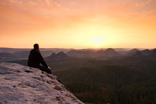 Adult tourist in black trousers, jacket sit on cliff's edge and looking to misty hilly valley bellow. Hikker relax and thinking alone in nature. 