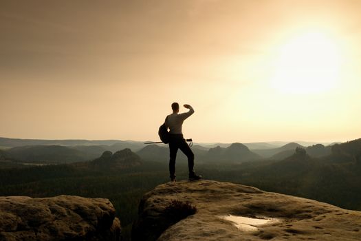 Tall backpacker with poles in hand. Sunny spring daybreak in rocky mountains. Hiker with big backpack stand on rocky view point above misty valley.