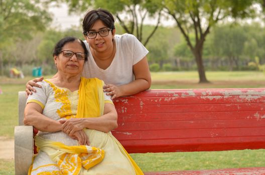 Happy looking young Indian woman with her mother sitting on a red bench in a park in New Delhi, India. Concept Mother's day