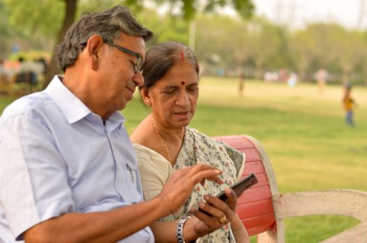 Portrait of senior couple sitting in park bench and looking at their smart phone and laughing in New Delhi, India with focus on the hands