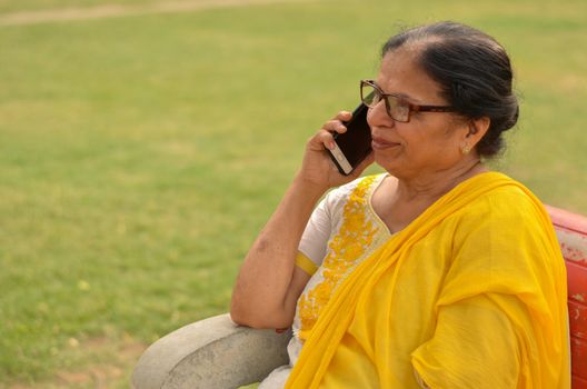 Side view Senior Indian woman speaking on her smart phone, sitting on a red bench in a park in New Delhi. Concept shot showcasing technology adoption by senior citizens. Digital India