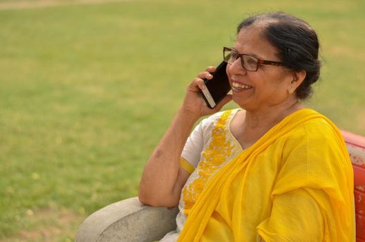 Side view Senior Indian woman speaking on her smart phone, sitting on a red bench in a park in New Delhi. Concept shot showcasing technology adoption by senior citizens. Digital India