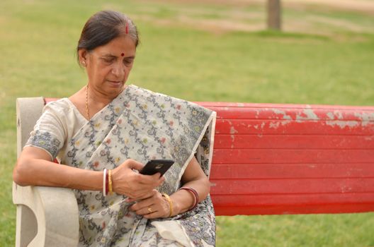 Senior Indian Bengali woman working on a mobile phone, experimenting with technology on a red park bench in an outdoor setting during summers wearing saree in New Delhi, India