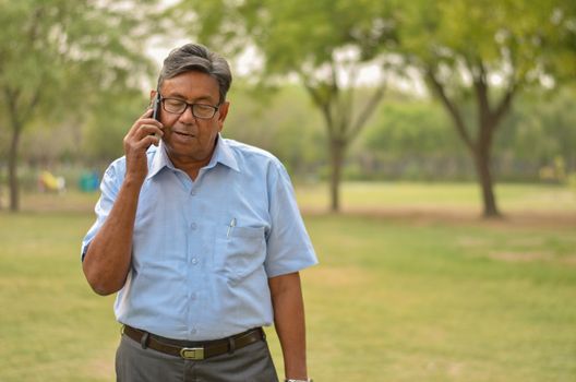 Senior businessman or working professional Indian man wearing a half sleeve shirt and trousers standing in a park speaking on his smart phone in Delhi. Concept digital literacy with senior citizens