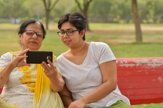 Young Indian girl with her old Indian mother looking at the mobile phone and busy talking selfie on a red bench in a park in New Delhi, India