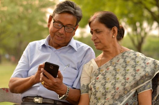 Portrait of senior couple sitting in park bench and looking at their smart phone and laughing in New Delhi, India with focus on the hands