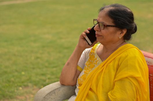 Side view Senior Indian woman speaking on her smart phone, sitting on a red bench in a park in New Delhi. Concept shot showcasing technology adoption by senior citizens. Digital India