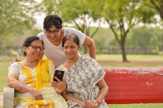 A young Indian girl with mother and mother in law - senior graceful women gossiping and smiling at the mobile phone screen while sitting on a red park bench in New Delhi, India. Concept Mother's day