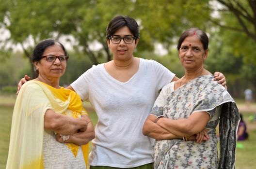 Portrait of two senior Indian women women standing with their daughter / daughter-in-law in a park wearing saree and salwar kamiz with crossed / folded hands during winters in New Delhi, India