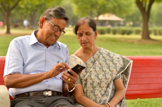 Portrait of senior couple sitting in park bench and looking at their smart phone and laughing in New Delhi, India with focus on the hands