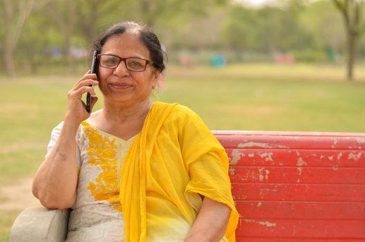 Senior Indian woman speaking on her smart phone, sitting on a red bench in a park in New Delhi. Concept shot showcasing technology adoption by senior citizens. Digital India