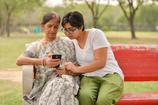 Young Indian girl with an old Indian Bengali woman looking at the mobile phone and busy talking sitting on a red bench in a park in New Delhi, India